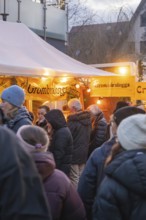 People stand at a brightly lit stand at a Christmas market, wrapped in warm clothes, Aidlingen