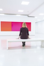 A woman sits in a modern, white art gallery and looks at a large, colorful painting, Open Horizons