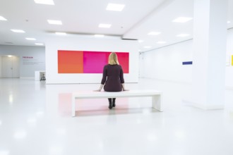 A woman sits alone on a bench in a bright white art gallery, Open Horizons exhibition, Schauwerk