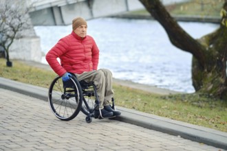 A man in a wheelchair moves along the river in the park taking a daily walk, wearing a red jacket
