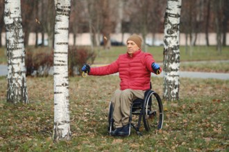 A senior man in wheelchair wearing red down jacket and gloves stretches arms outdoors in a grassy