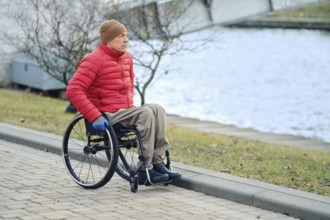 A man in a wheelchair stopped by a canal in a city park and looks at the water. He wears a red down