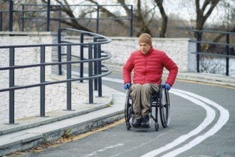 An elderly man wearing a red down jacket and a hat moves steadily in his wheelchair along a curvy