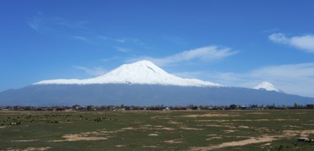 Snow-covered mountain range above a small village surrounded by green fields under a blue sky,