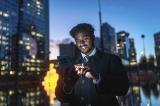 Young man standing outdoors at night, smiling while interacting with a smartphone, reflecting