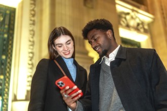 Diverse couple standing close, looking at a smartphone screen, smiling and connecting while sharing