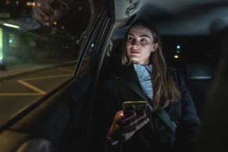 Woman travelling in the back seat of a car at night, holding a smartphone and looking out the
