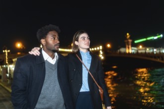 Young diverse couple strolling along a river promenade at night, city lights reflecting on the
