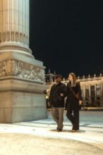 Interracial couple strolling arm in arm past an illuminated historic monument at night, sharing a