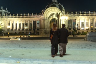 Couple walking arm in arm beneath the illuminated facades of petit palais at night, enjoying a