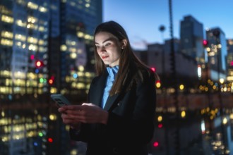 Young woman standing outdoors at night, using her smartphone with a glowing screen, surrounded by