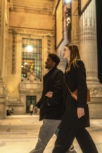 Diverse couple walking arm in arm on an illuminated city street at night, exploring urban