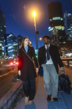 Diverse business couple walking with luggage along a downtown sidewalk at night, passing