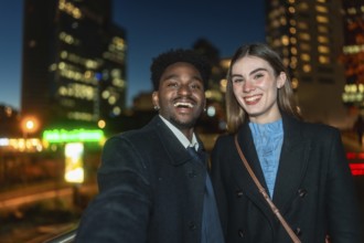 Diverse young man and woman smiling happily while taking a selfie together outdoors at night, with