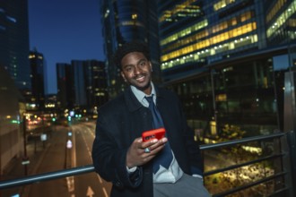 Young black businessman standing on a bridge, holding a modern smartphone with a red case, smiling