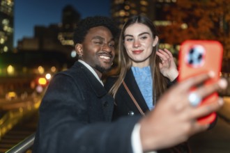 Diverse young couple on a city street at night, smiling and taking a candid selfie with a