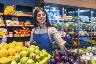 Greengrocer woman smiling as she arranges vibrant fresh plums and assorted produce at a market