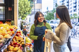 Smiling greengrocer wearing an apron and gloves receiving a female customer holding a reusable net