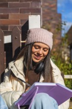 Young woman in a beanie and headphones writing in a notebook outdoors on a sunny day, smiling and