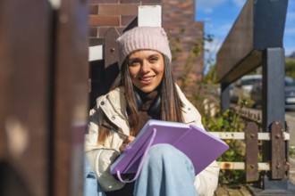 Young woman in beanie and headphones smiles while sketching in a notebook on a sunny urban street,