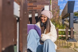 Young woman sitting on the sidewalk enjoying a good book, dressed in a warm winter coat and beanie,