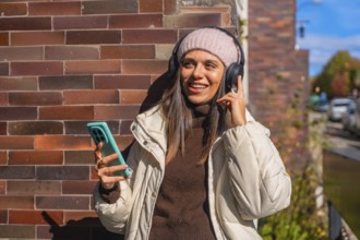 Young woman smiling while enjoying music through her headphones, holding a smartphone outdoors