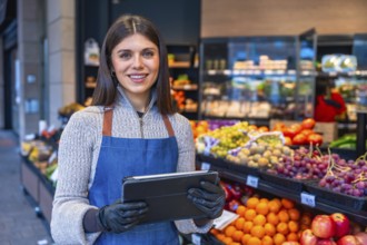 Greengrocer woman wearing an apron and gloves, standing in a small business produce shop, holding a
