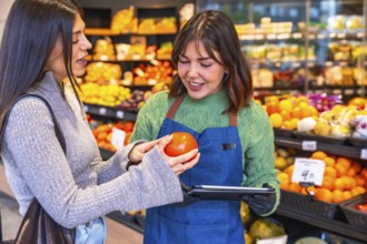 Grocery store employee in an apron holding a tablet and helping a female customer examine a fresh