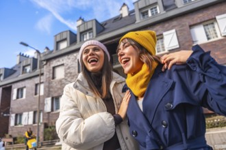 Two cheerful young women friends enjoying a sunny city day, laughing wholeheartedly while wearing