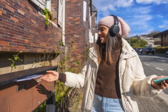 Young woman smiling, wearing a beanie and headphones, mailing a letter into an outdoor mailbox on