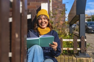 Young woman smiling in a cozy beanie and scarf, sitting on a city street enjoying a sunny autumn
