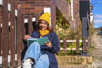 Woman wearing a yellow beanie and blue coat sitting on steps, enjoying a book in the warm sunlight