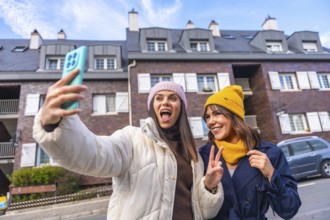 Two cheerful young women take a sunny winter selfie on a city street, bundled in beanies and