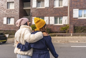 Two young women in warm winter clothes walk arm in arm down a city street, smiling and enjoying a