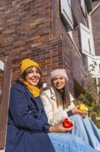 Two friends in warm winter clothes sit against a brick wall, smiling and sharing red and yellow