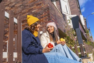 Two young women sit on outdoor steps by a brick wall under blue sky, smiling and chatting while