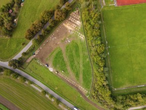 Aerial view of a field with construction next to a sports field, surrounded by trees, construction