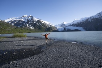 Young man jumping on a pebble beach on a glacial lake, Snowy Mountains and Portage Glacier on