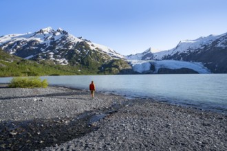 Young man on a pebble beach on a glacial lake, snowy mountains and Portage Glacier glacier on