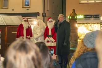 Two Santa Clauses stand with a woman and a speaker in front of an audience at a Christmas market,