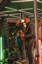 Two musicians on a stage at night playing guitars surrounded by festive lighting, Christmas market