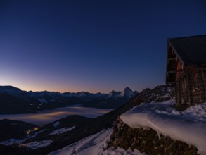 Alpine panorama in winter with Watzmann and alpine hut under starry sky at dawn, fog in the valley,