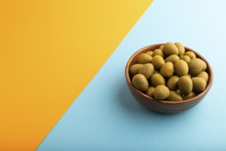 Fresh green olives in wooden bowl on blue and orange pastel paper background. side view, copy space