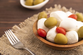 White Mozzarella cheese, with tomatoes and olives in wooden bowl on brown wooden background and