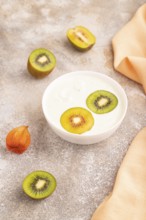 Yogurt with kiwi in white bowl on brown concrete background and orange linen textile, side view,