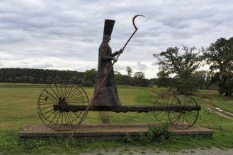 Sculpture in a meadow, traditional grim reaper, figure carrying a scythe, standing on cart with