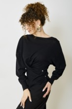 A stylish model with curly hair poses against a neutral backdrop. She wears black blouse with twist
