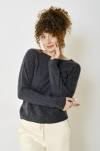 Cheerful woman in dark sweater and light pants standing against white wall with a playful