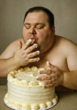 A very fat obese man sits in front of a buttercream cake and licks his fingers off with butter