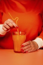 A woman in an orange sweater, stirring with a straw, orange juice in a glass, orange background